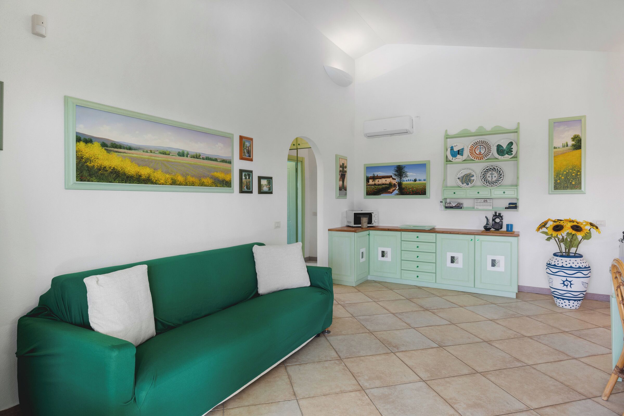 A room with a green sofa, a kitchen area with light green cabinets, and a large framed landscape painting on the wall.
