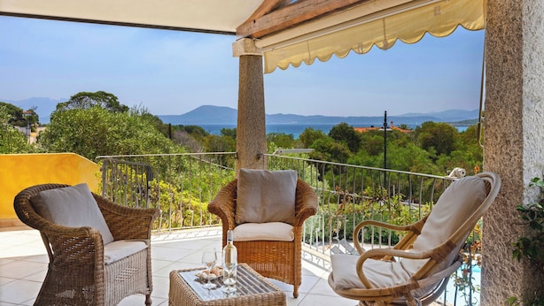Un balcon agrémenté de meubles en osier, une table avec une bouteille et des verres, et une vue panoramique sur la mer et les montagnes.