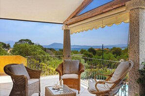 A balcony with wicker furniture, a table with a bottle and glasses, and a scenic view of the sea and mountains. - Sardegna è - Beach Apartments LE  (Pittulongu)