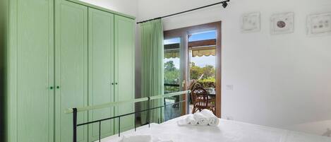 A bedroom with a bed, a green wardrobe, a balcony with a table and chairs, and a view of greenery.