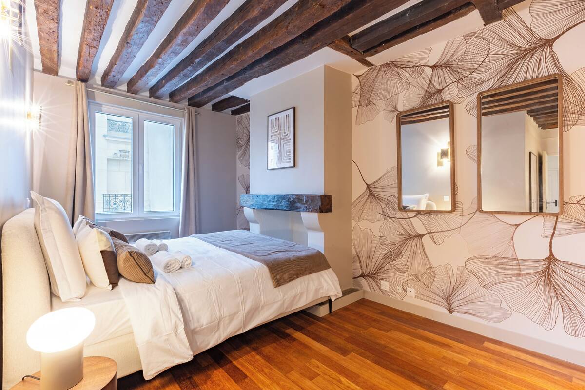 A bedroom with a wooden ceiling, a bed with white bedding, a bedside table with a lamp, and a wall with a leaf pattern.