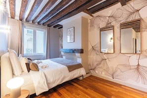 A bedroom with a wooden ceiling, a bed with white bedding, a bedside table with a lamp, and a wall with a leaf pattern. - Appartements rue des Vertus (Paris)
