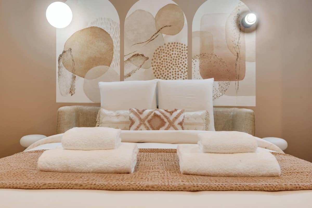 A neatly made bed with two towels, a textured rug, and a wall with abstract art.