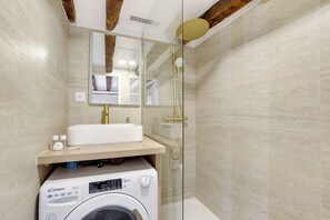 A modern bathroom with a glass-enclosed shower, a white sink, and a built-in washing machine. - Appartements rue des Vertus (Paris)