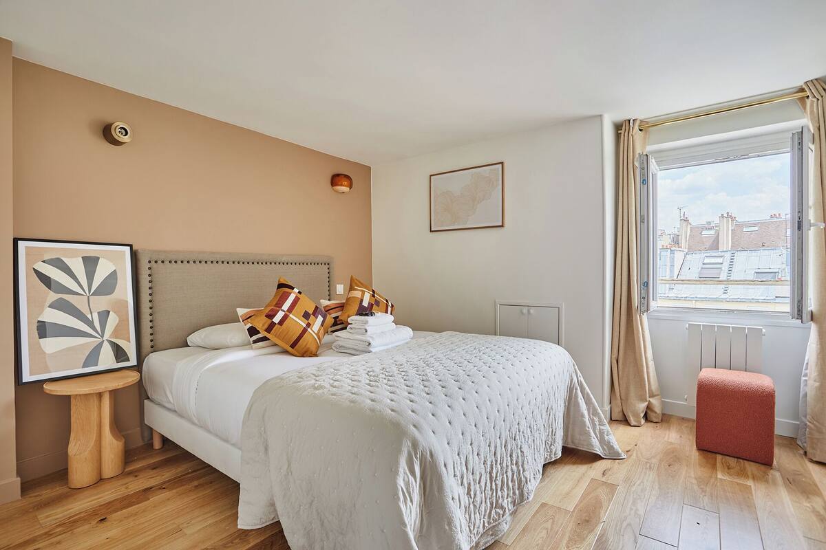A modern bedroom with a large bed, a small table, a radiator, and a window with curtains.