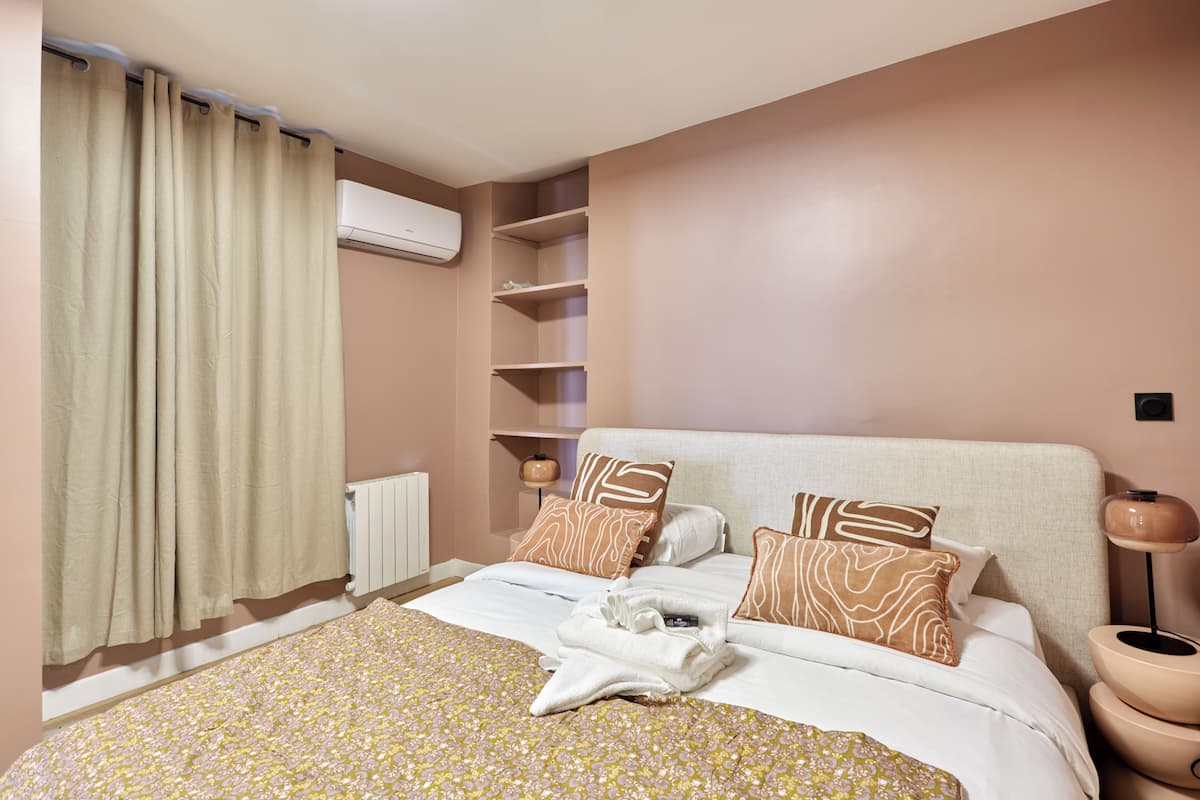 A hotel room with a bed, a radiator, a shelf unit, and a bedside table with a lamp.