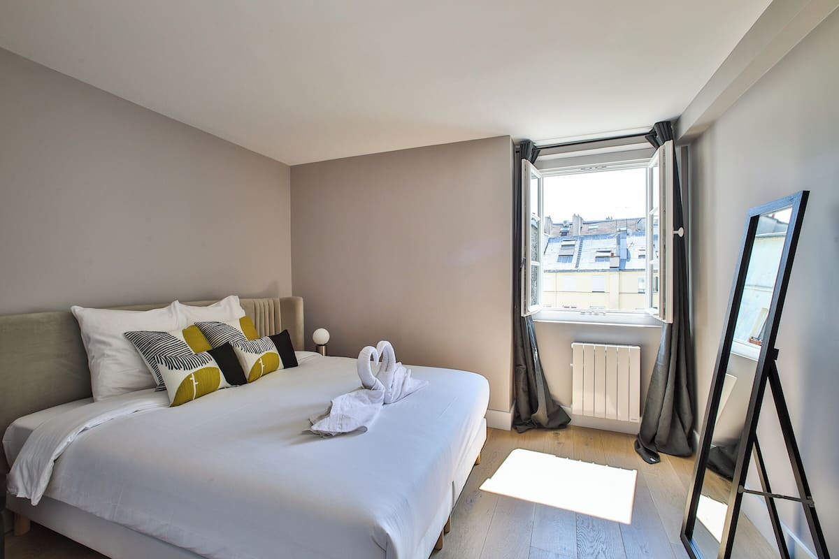 A hotel room with a bed, a window with curtains, and a standing mirror.