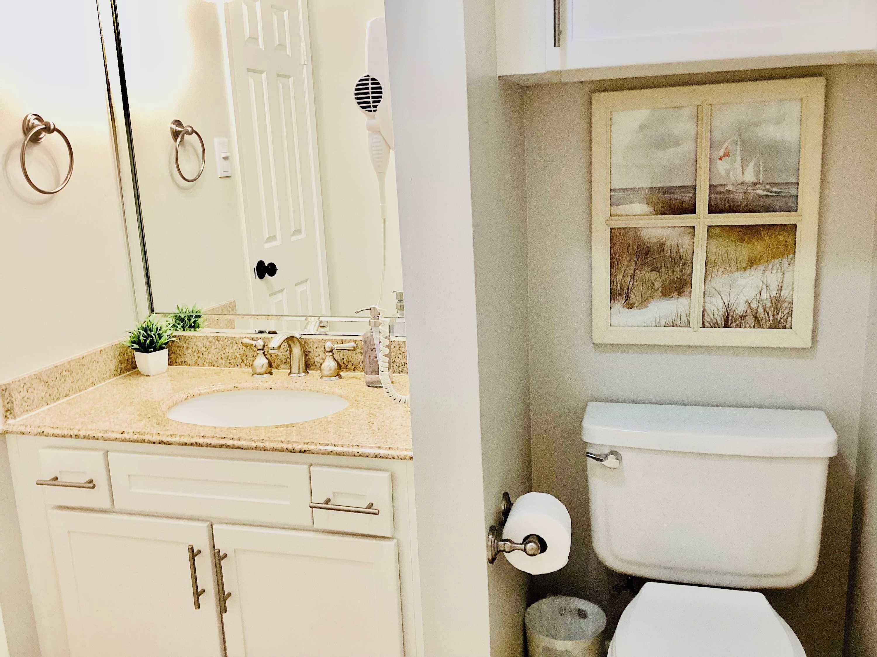 Baño con lavamanos, espejo e inodoro. Se ve una ventana pequeña con un cuadro sobre el inodoro.