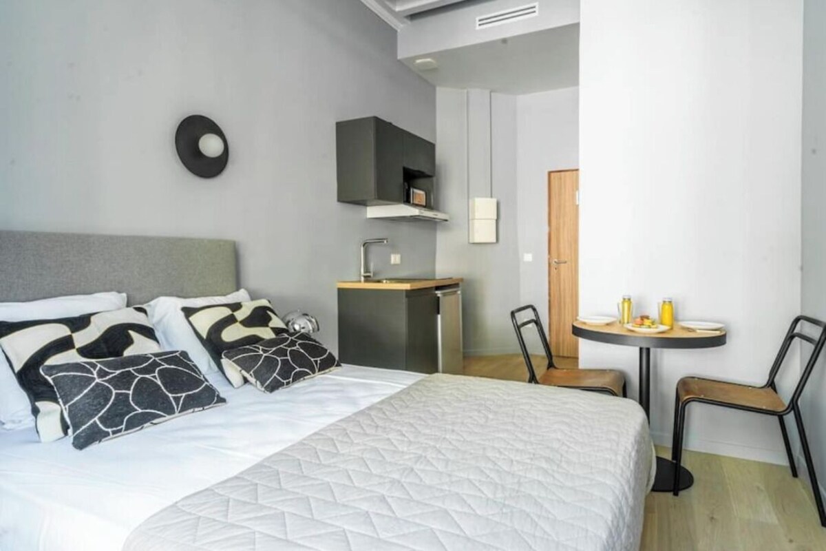Una habitación de hotel moderna con cama, una kitchenette pequeña, una mesa redonda con sillas y un espejo.
