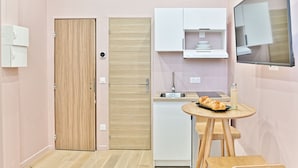 A compact kitchen with a wooden door, white cabinets, a sink, and a small dining table with a bottle and croissants. - Apparements Berthier (Paris)