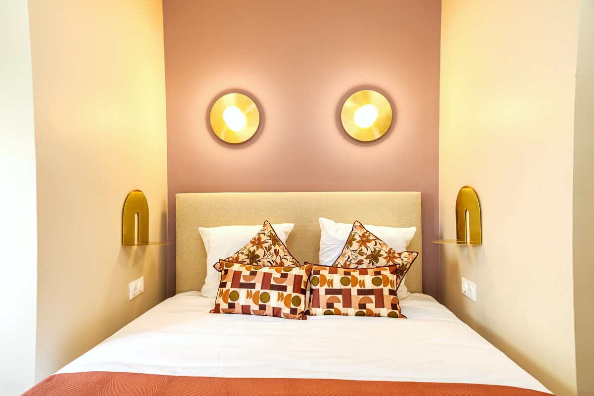 Una cama bien hecha con almohadones estampados, dos luces fijadas a la pared y lámparas de noche.