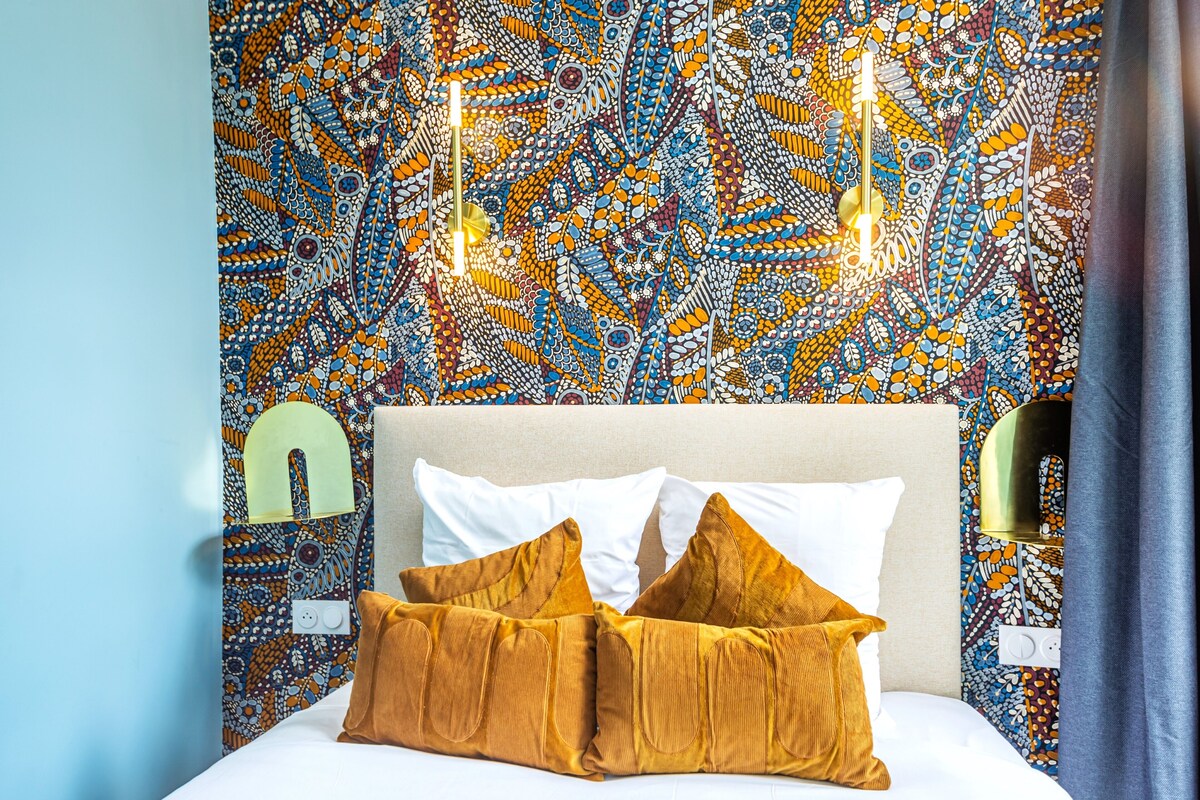 Una cama con almohadas blancas y mostaza, un cabecero color beige y una pared estampada con una lámpara.