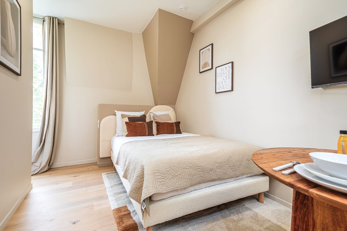 Un dormitorio con cama, una mesita de noche con un cuenco y una botella, un televisor montado en la pared y una ventana con cortinas.