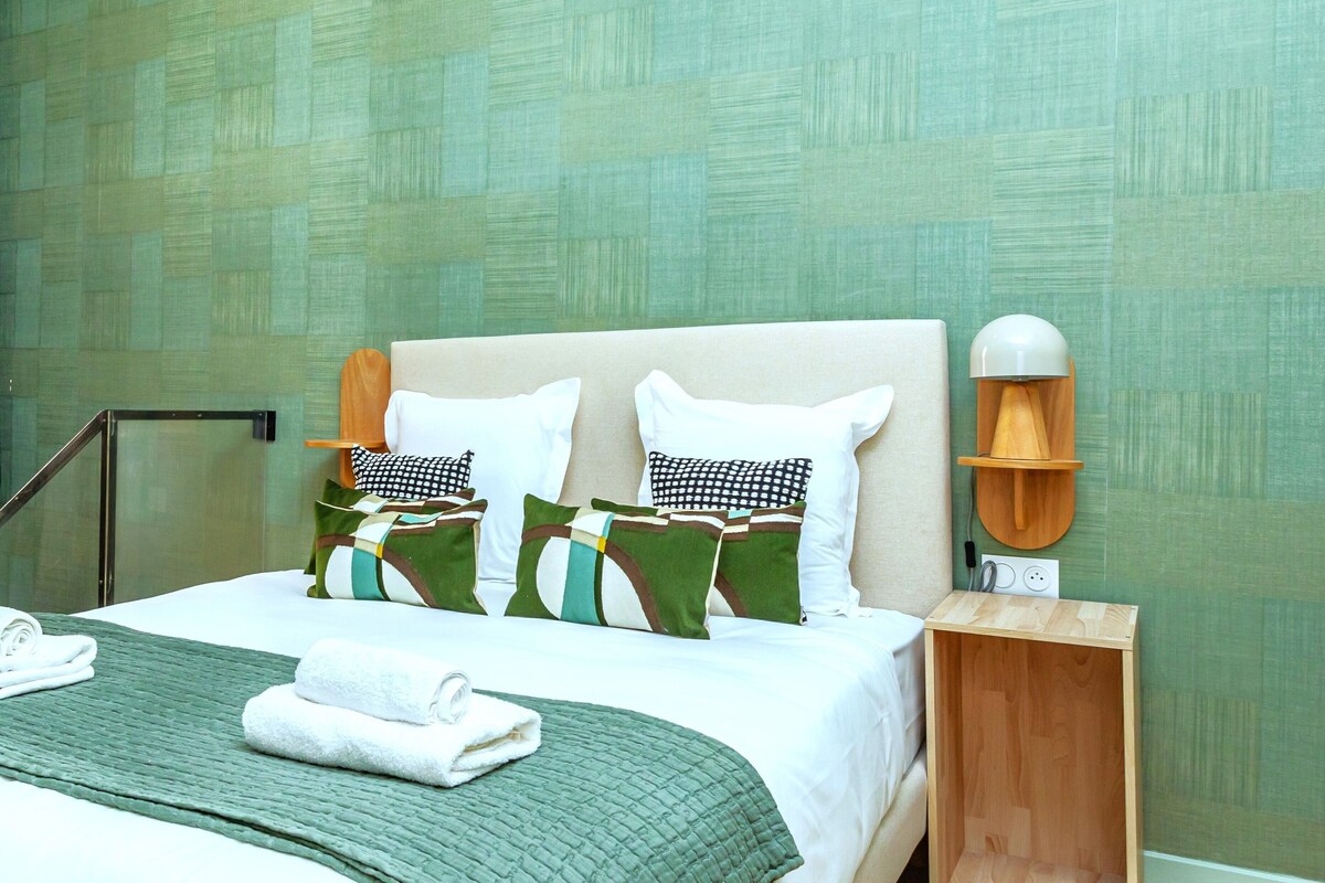 Una cama bien hecha con una manta verde y almohadas blancas, una mesita de noche de madera con una lámpara, y una pared con un diseño texturizado.