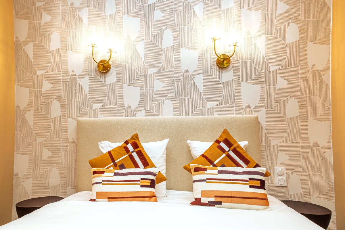 Una habitación de hotel con papel tapiz estampado, un cabecero color beige y dos mesitas de noche con lámparas.
