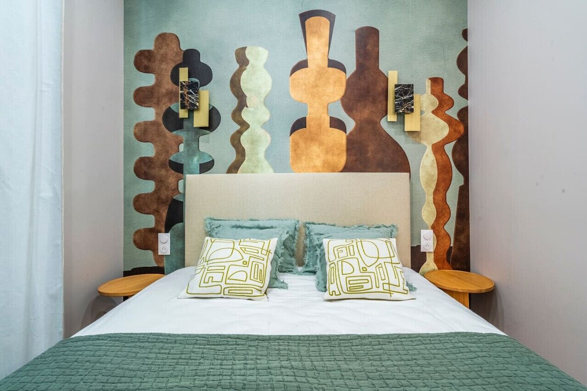 Una cama con colcha verde, dos almohadas con diseños geométricos y una pared con arte abstracto.