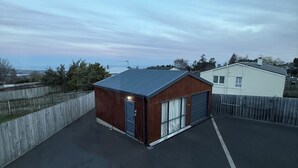 A small, single-story building with a dark roof and a wooden facade, situated in a fenced area with a paved driveway. - A private mini house with own T&B, mini kitchen and entertainment area! (Dunedin)