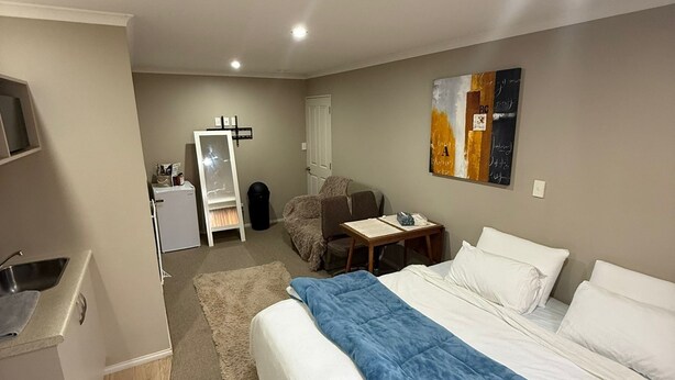 A hotel room with a bed, a desk, a chair, a small table, a refrigerator, a sink, and a painting on the wall. - A private mini house with own T&B, mini kitchen and entertainment area! (Dunedin)