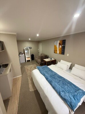 A hotel room with a bed, a desk, a chair, a small table, a refrigerator, a sink, and a painting on the wall. - A private mini house with own T&B, mini kitchen and entertainment area! (Dunedin)