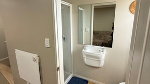 A bathroom with a white sink, a mirror, and a shower. There is a blue bath mat on the floor. - A private mini house with own T&B, mini kitchen and entertainment area! (Dunedin)