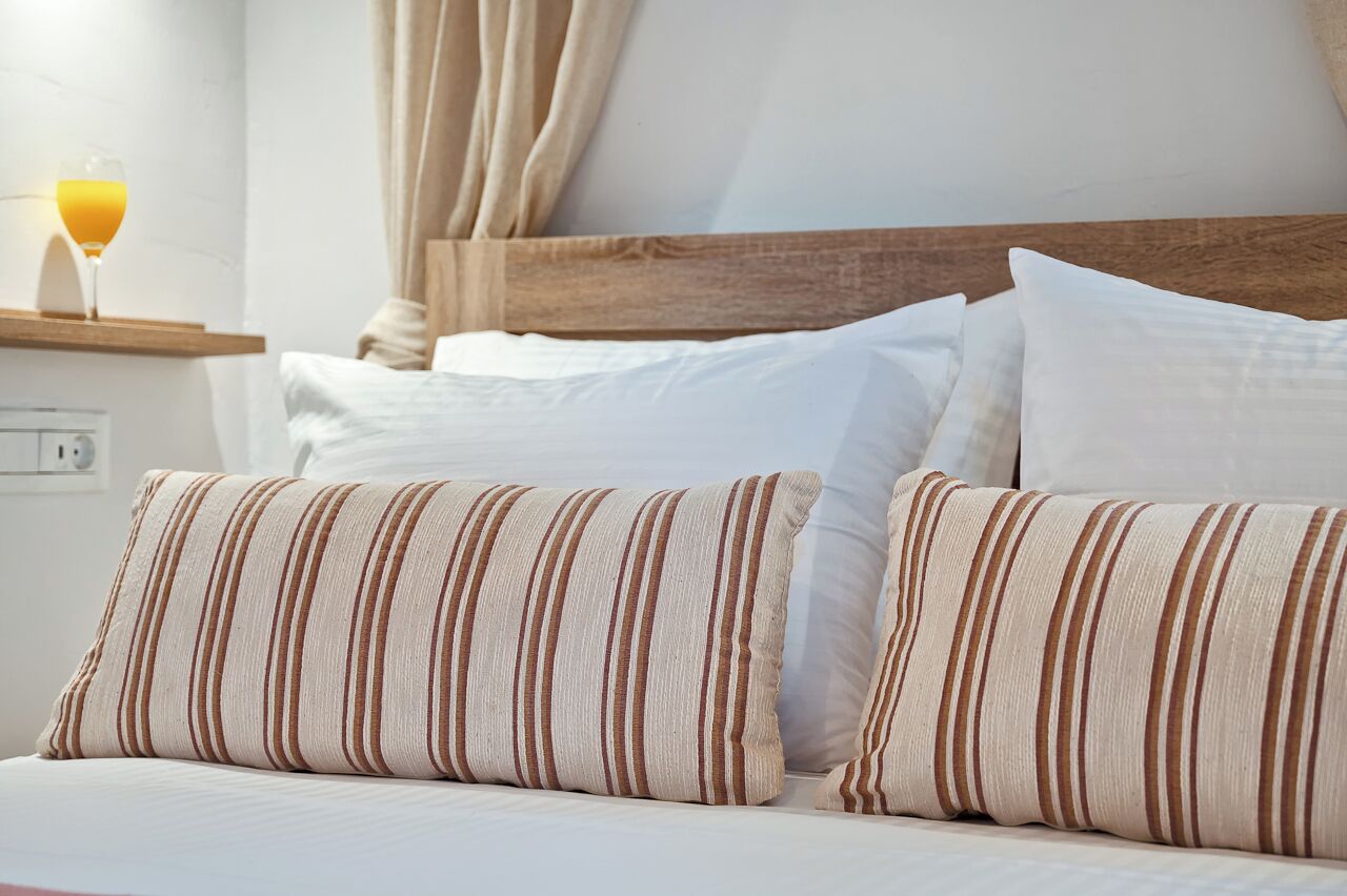 Una cama bien hecha con almohadas a rayas, un cabecero de madera y una mesita de noche con un vaso de jugo de naranja.