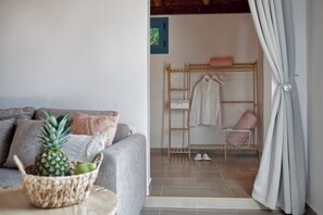 Una sala de estar acogedora con un sofá gris, una cesta tejida con una piña y un perchero de madera con ropa y zapatos.