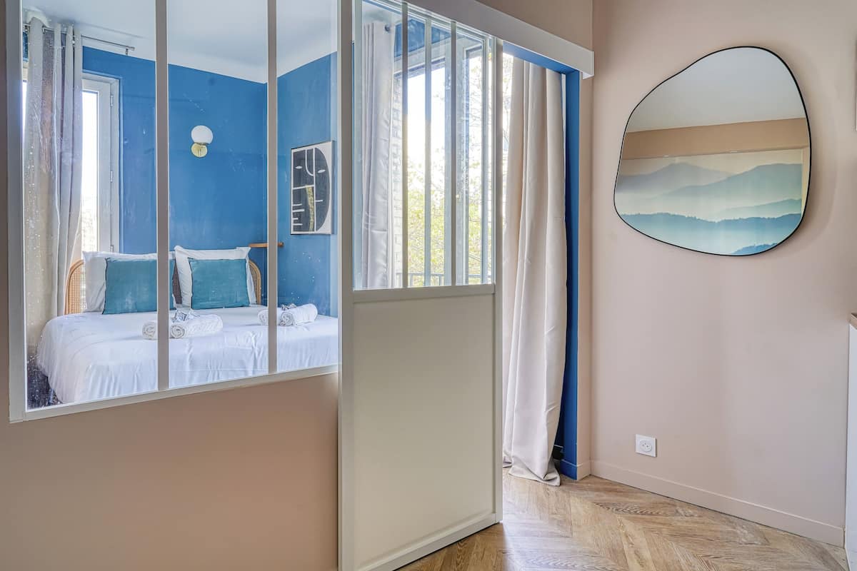 Un dormitorio con un ventanal grande, una cama con ropa de cama blanca, un espejo montado en la pared y un cuadro de montañas.