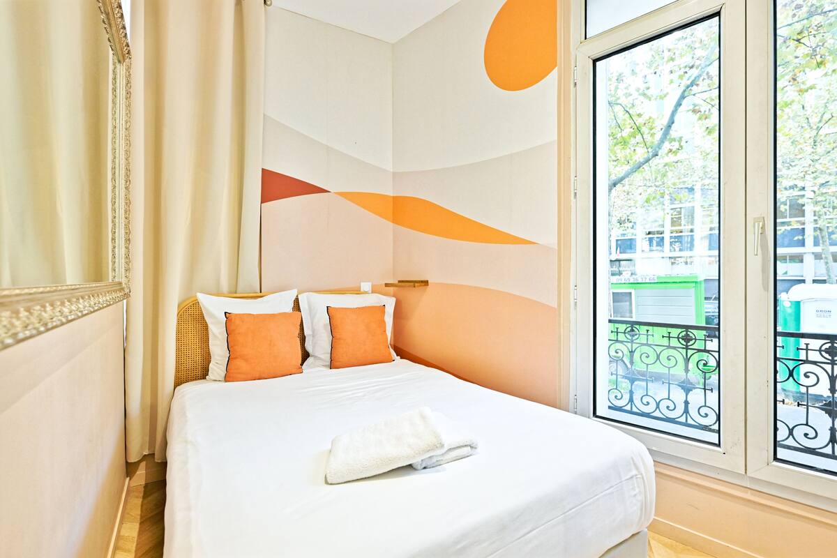 Una cama bien hecha con ropa de cama blanca y naranja, un ventanal con vista a una casa verde y una pared decorativa con formas abstractas.