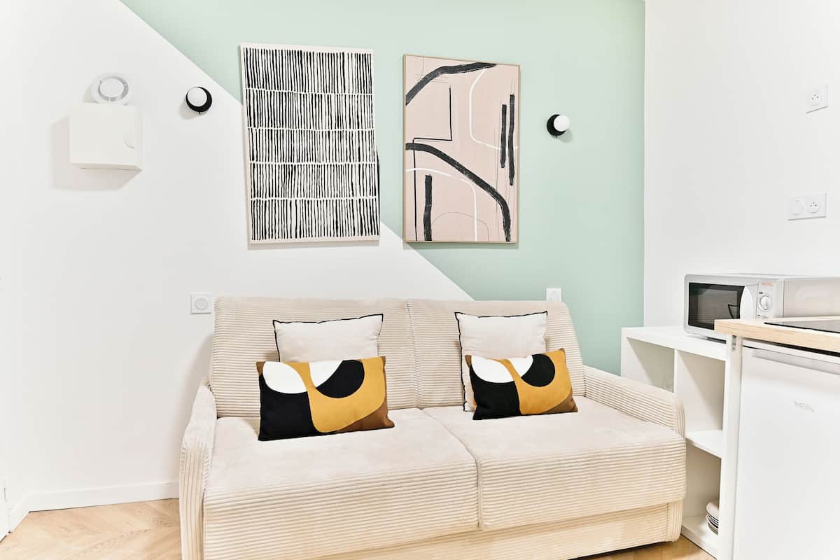 Una sala de estar moderna con un sofá color beige, dos cuadros abstractos, un calentador blanco montado en la pared y un mueble blanco con un microondas.