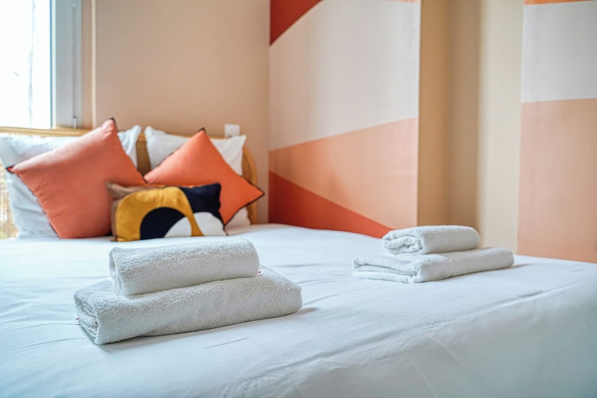 Una cama bien hecha con sábanas blancas y toallas dobladas, un colorido arreglo de almohadas y un diseño geométrico en la pared.