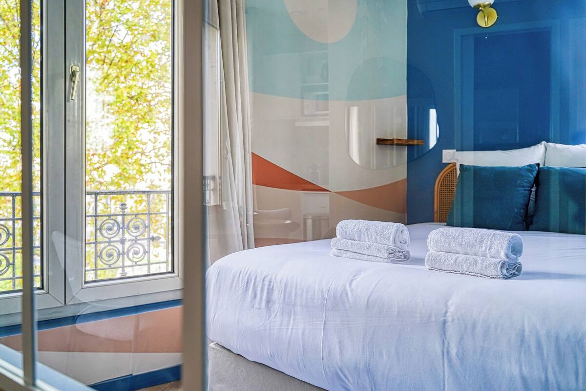 Habitación de hotel con una cama grande, dos toallas enrolladas, una pared azul y vistas a árboles a través de una puerta de cristal.