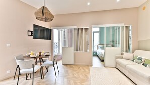 A modern living room with a sofa, dining table, and chairs. There is a television mounted on the wall and a window with curtains. - Appartements avenue Gourgaud (Paris)