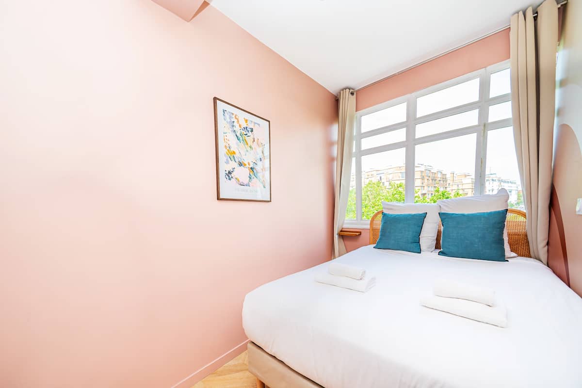 Un dormitorio con una cama grande, una ventana con cortinas, un cuadro en la pared y vistas a edificios.