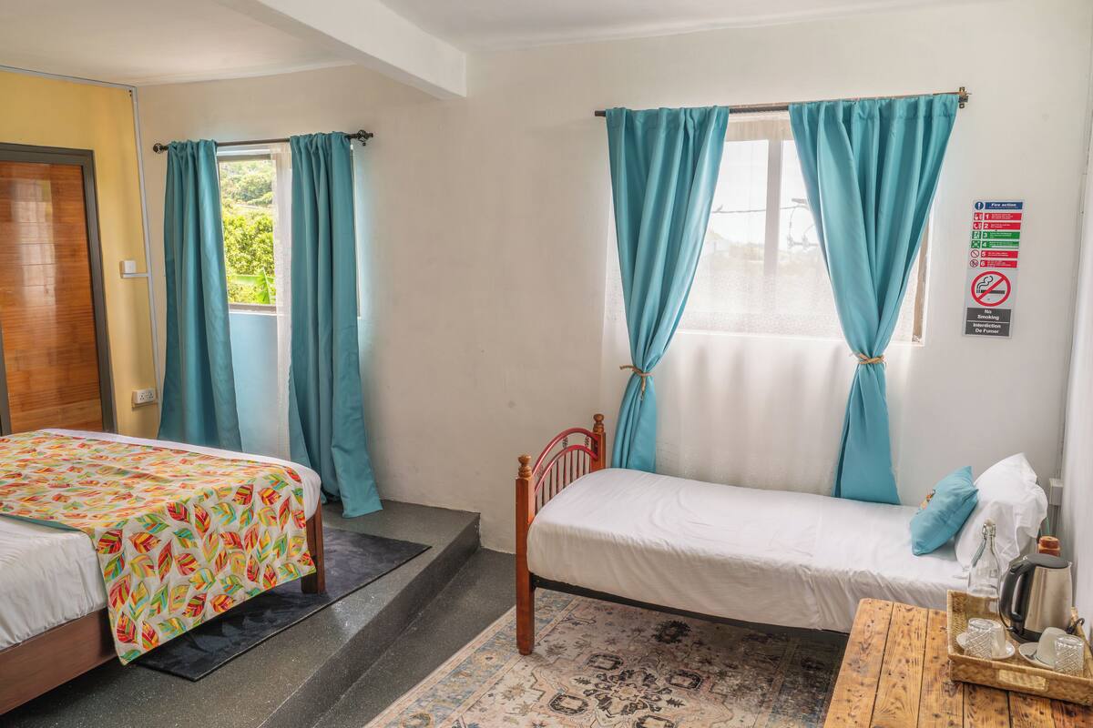 Una habitación con dos camas, una mesa de madera con una bandeja, un ventanal con cortinas y un letrero en la pared.