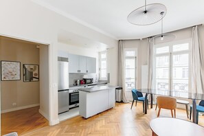 Una cocina moderna con una isla central, comedor con mesa y sillas de vidrio, y ventanas amplias con cortinas.