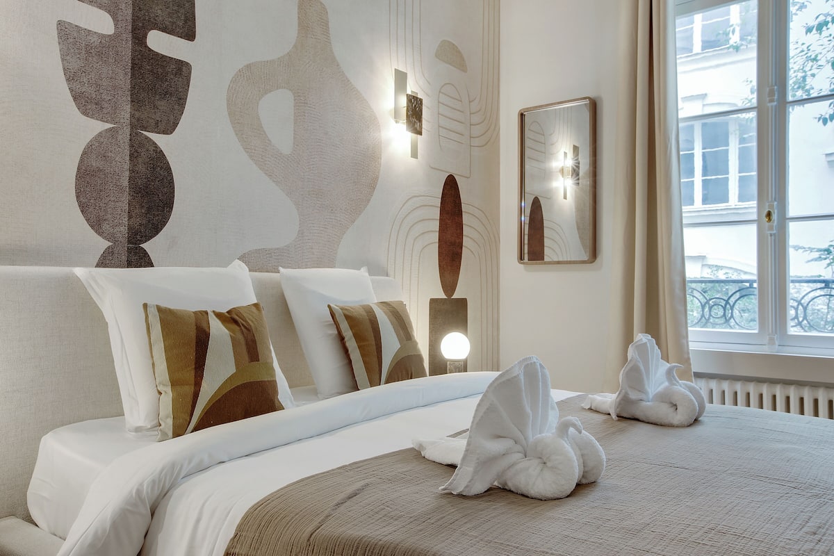 A hotel room with a neatly made bed, a large abstract wall art piece, and a window with a view of buildings.