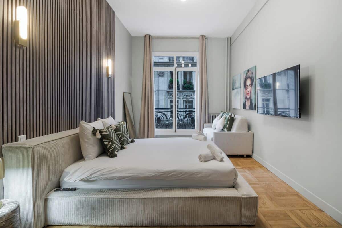 A modern bedroom with a large bed, a sofa, a TV, and a balcony with a view of buildings.