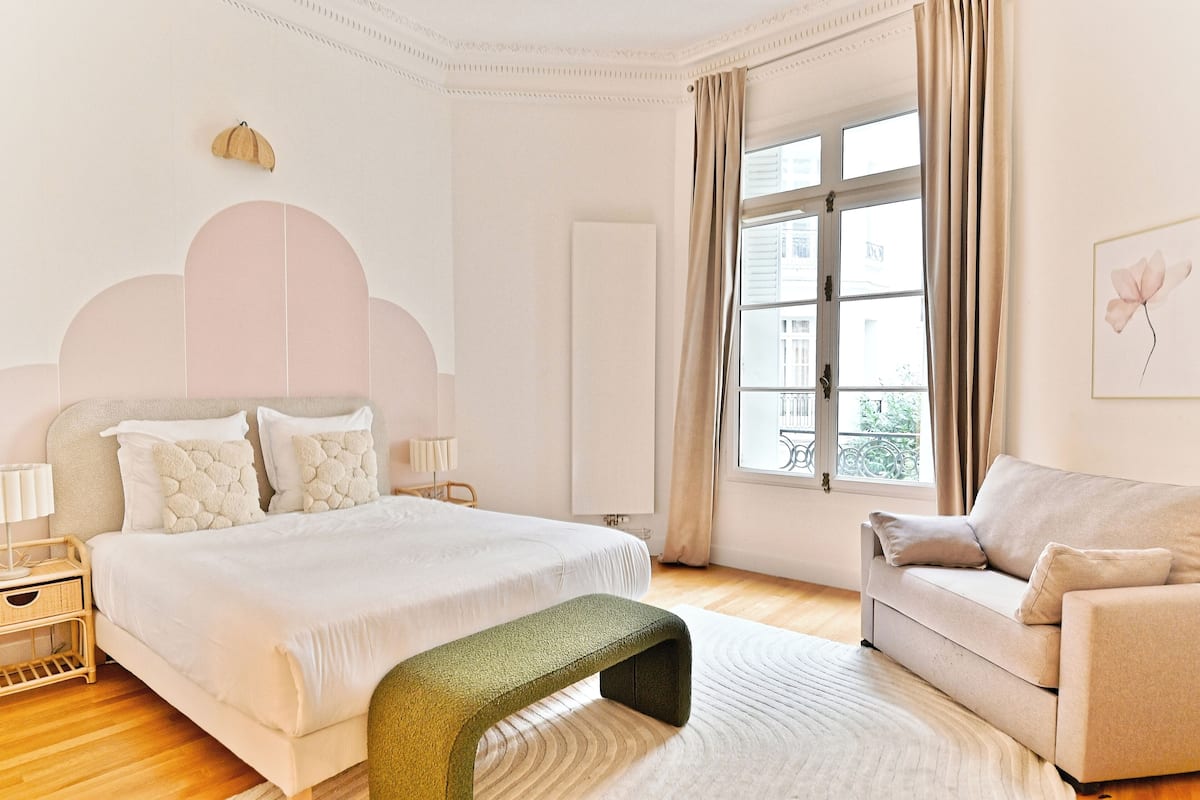 A bedroom with a bed, a bench, a sofa, a window, and a wall-mounted lamp.