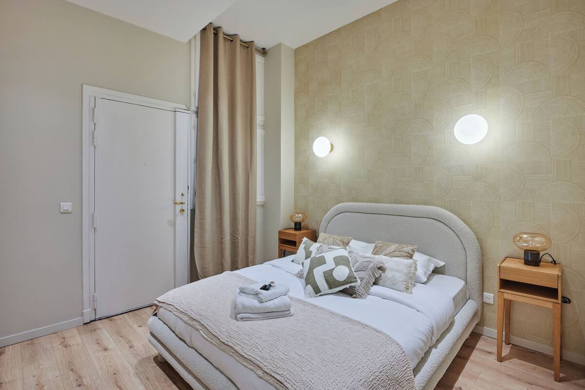 A neatly made bed with a white comforter and pillows, a wooden nightstand with a lamp, and a wall-mounted light fixture.