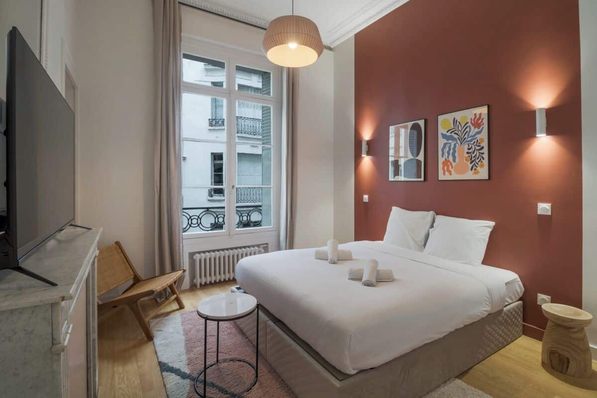 A hotel room with a bed, a small round table, a chair, a TV, and a window with a view of buildings.