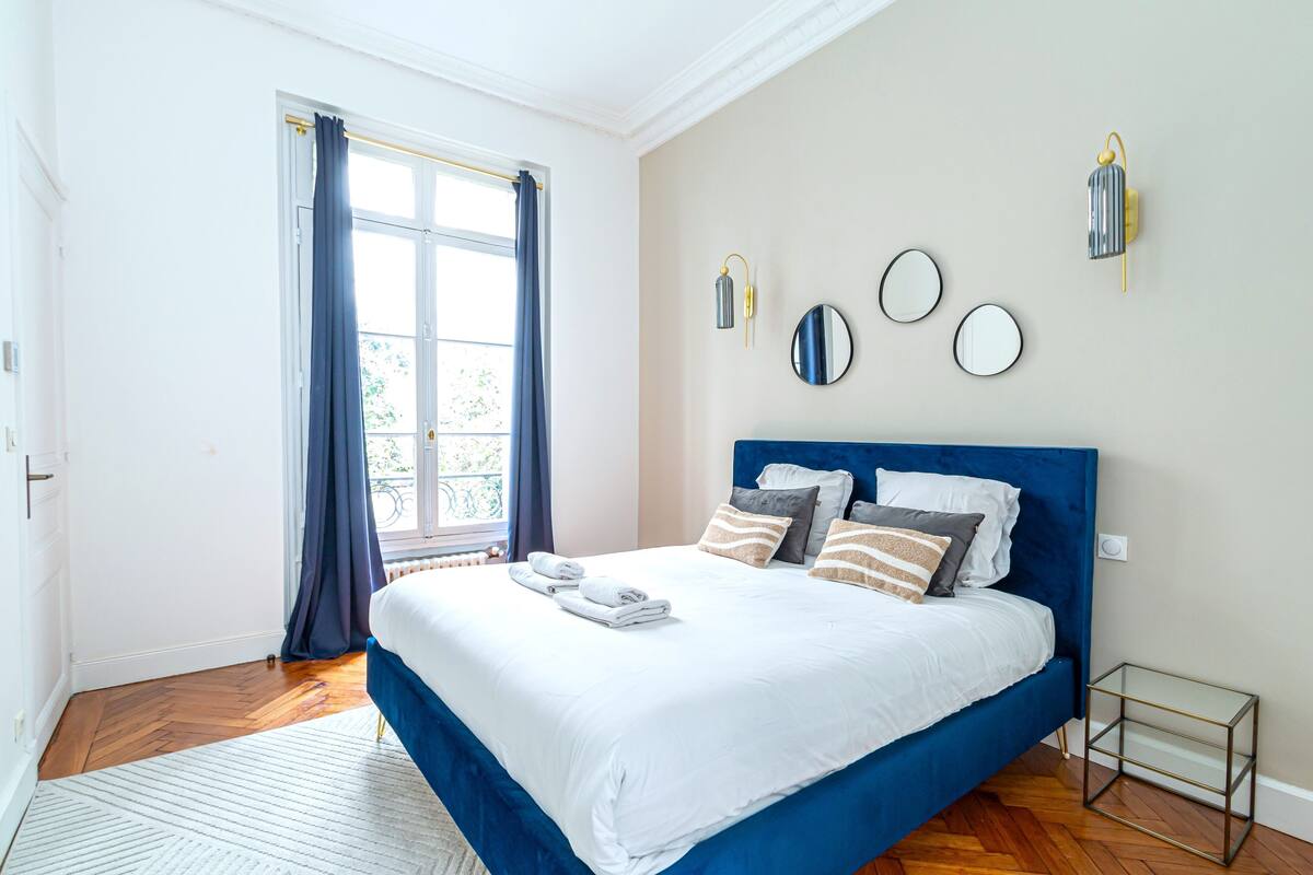 A bedroom with a large bed, a window with curtains, a radiator, and wall-mounted mirrors.