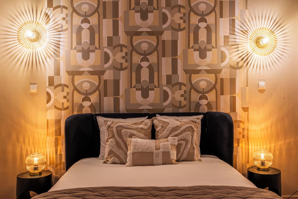 A modern bedroom with a geometric patterned wall, a dark headboard, and two bedside lamps.