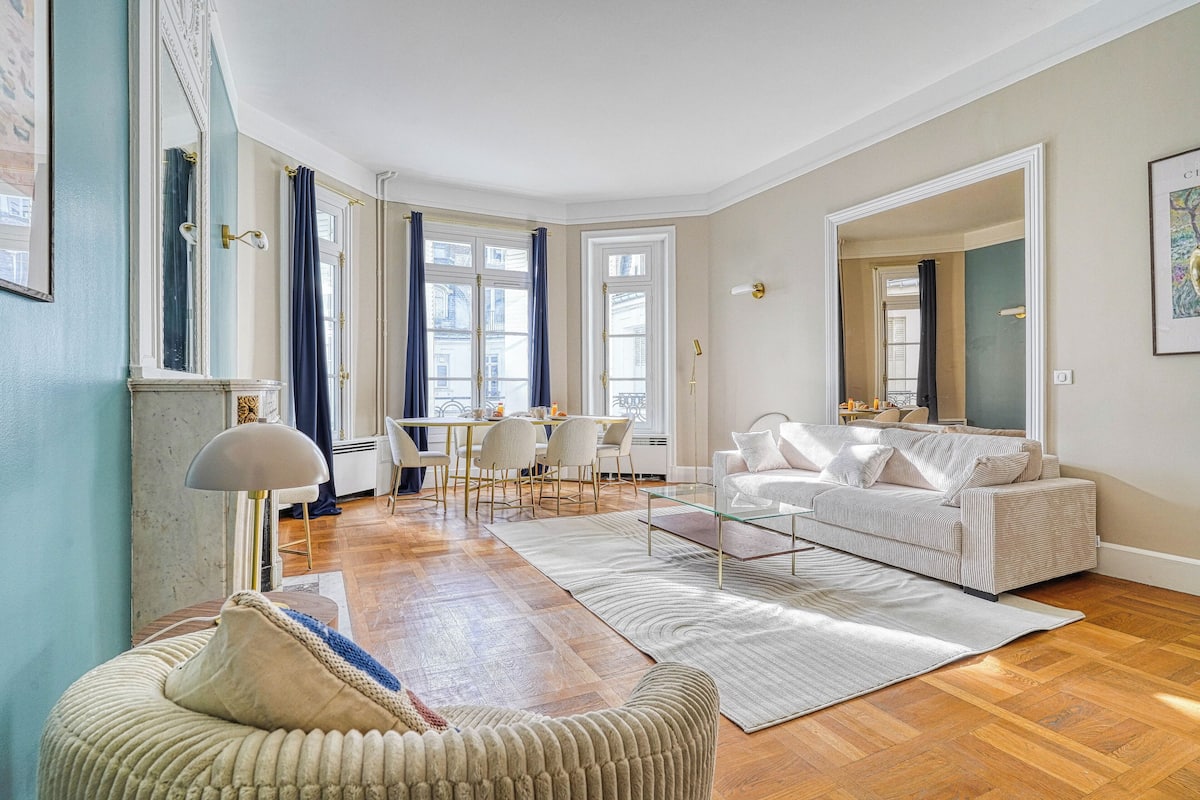 A spacious living room with a large window, a sofa, a coffee table, and a dining area with chairs and a table.
