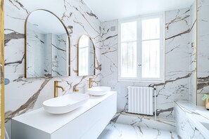 Un baño con paredes de mármol, un tocador blanco con dos lavamanos, accesorios dorados y un espejo grande.