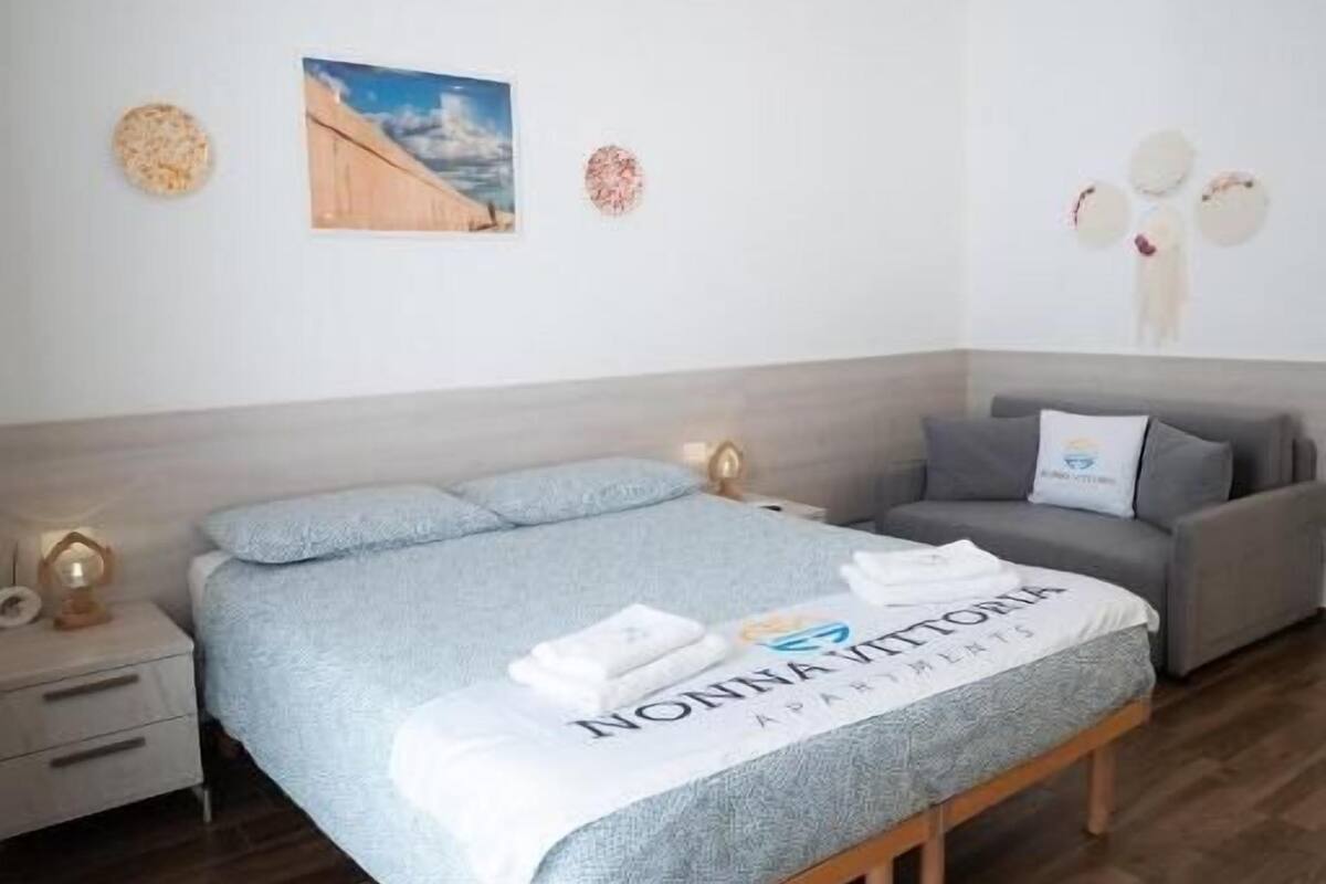 Un dormitorio ordenado con cama, mesita de noche, sofá y una lámpara fijada a la pared.