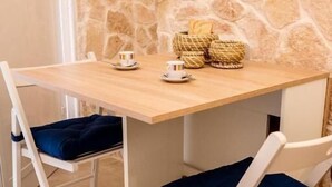 A small dining area with a stone wall, a wooden table, two white chairs with blue cushions, and a basket of bread on the table. - Nonna Vittoria Apartments (Mola di Bari)