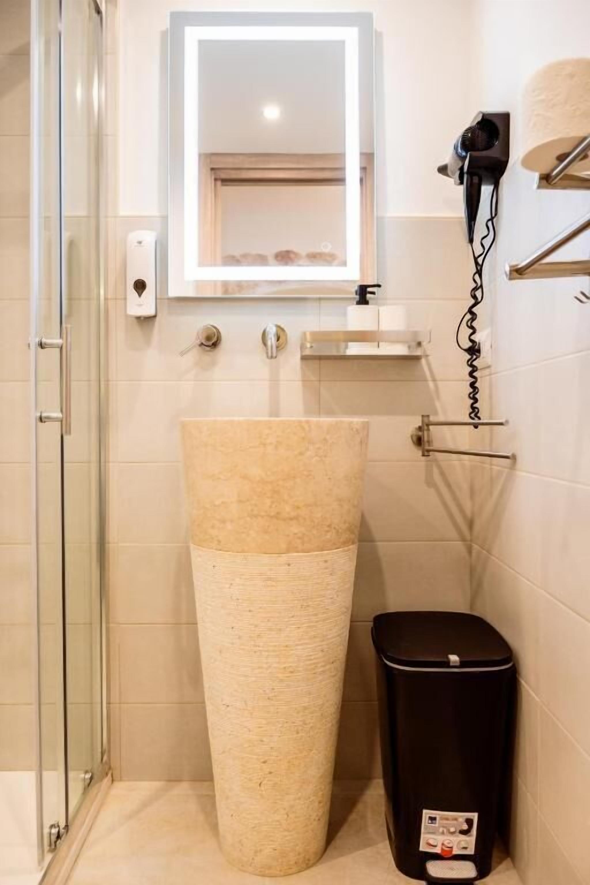 Un baño con un lavamanos grande color beige, un bote de basura negro, un secador de pelo en un soporte de pared y una cabina de ducha con paredes de vidrio.