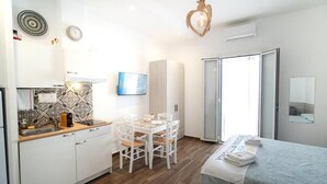 A compact room with a kitchenette, a dining table with chairs, a bed with a folded towel, and a sliding glass door leading to a balcony. - Nonna Vittoria Apartments (Mola di Bari)