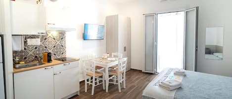 A compact room with a kitchenette, a dining table with chairs, a bed with a folded towel, and a sliding glass door leading to a balcony.