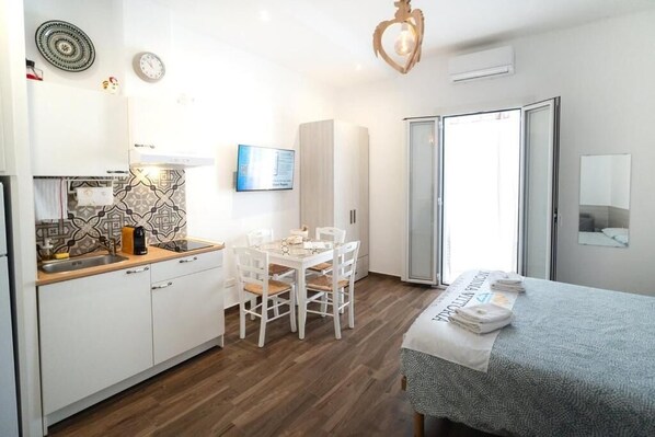A compact room with a kitchenette, a dining table with chairs, a bed with a folded towel, and a sliding glass door leading to a balcony. - Nonna Vittoria Apartments (Mola di Bari)